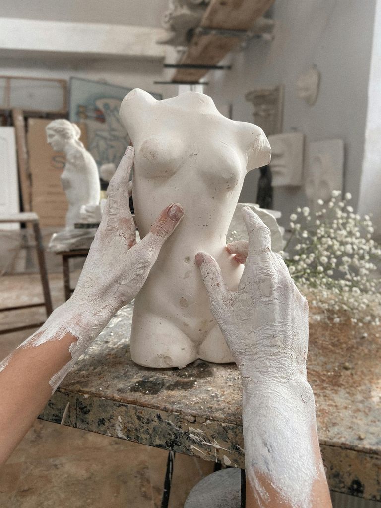 Unrecognizable talented artisan with dirty hands making clay female torso while sitting at wooden workbench with branch in modern workshop